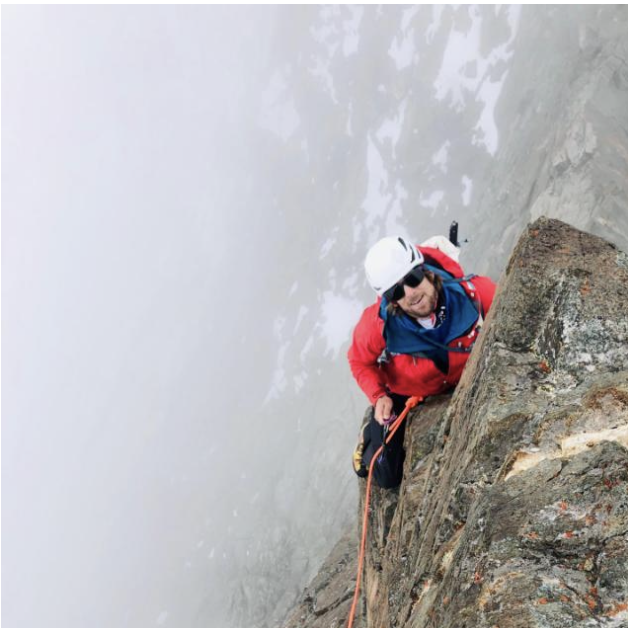 Quentin Cheseaux en veste rouge et casque, souriant en arrivant au sommet d'une arête rocheuse au-dessus des nuages